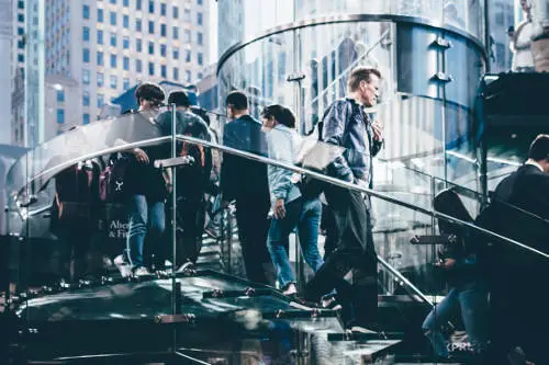 A group of people walks up and down a glass spiral staircase in a modern urban setting. The image features tall buildings and reflective surfaces, capturing the bustling city atmosphere. Amidst this scene, a man in a leather jacket descends the stairs, perhaps pondering the tax advantages of permanent life insurance.